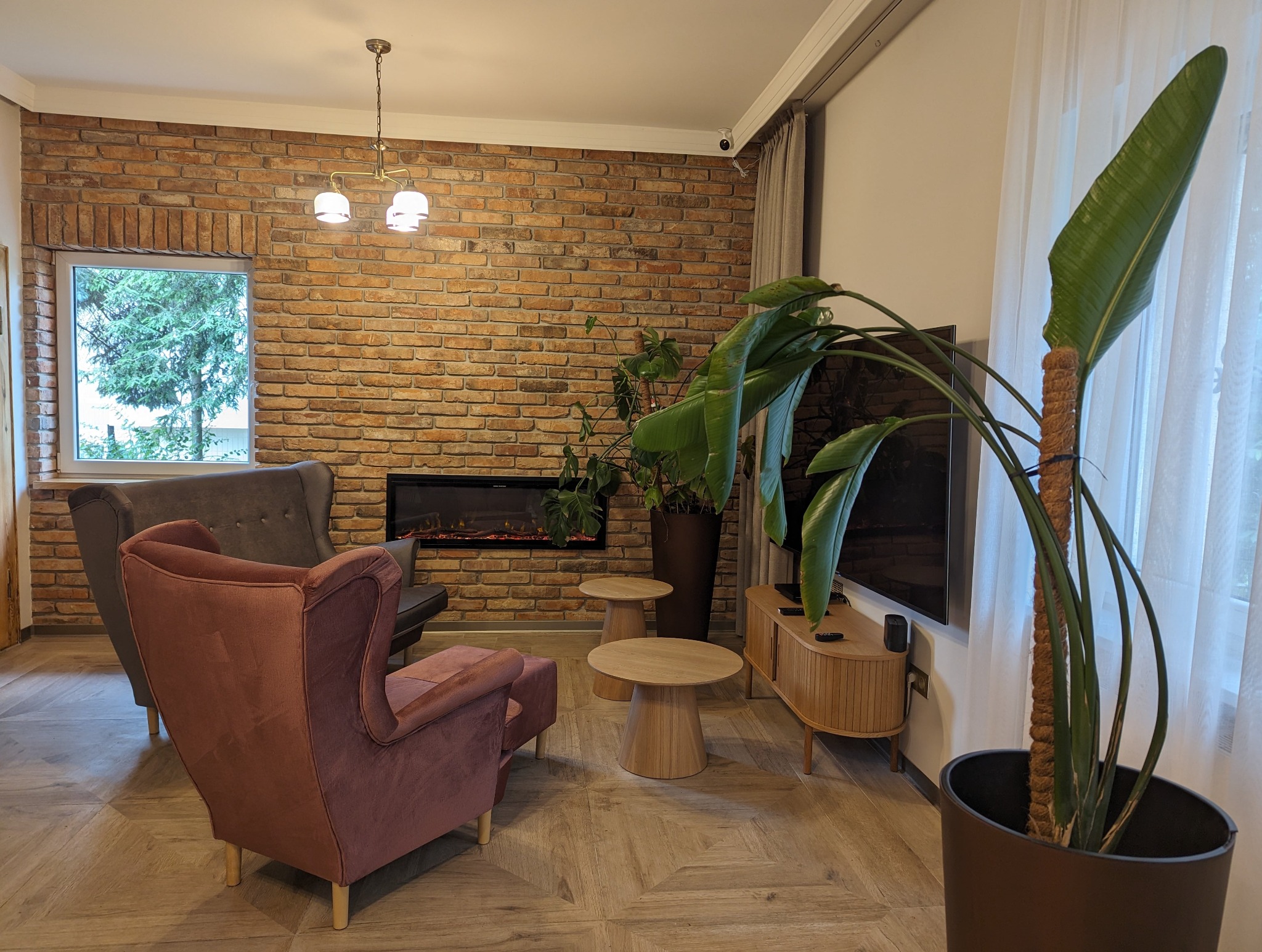 Salon z ceglaną ścianą, dwoma fotelami w odcieniach brązu, elektrycznym kominkiem i drewnianymi stolikami kawowymi na parkiecie ułożonym we wzór jodełki.