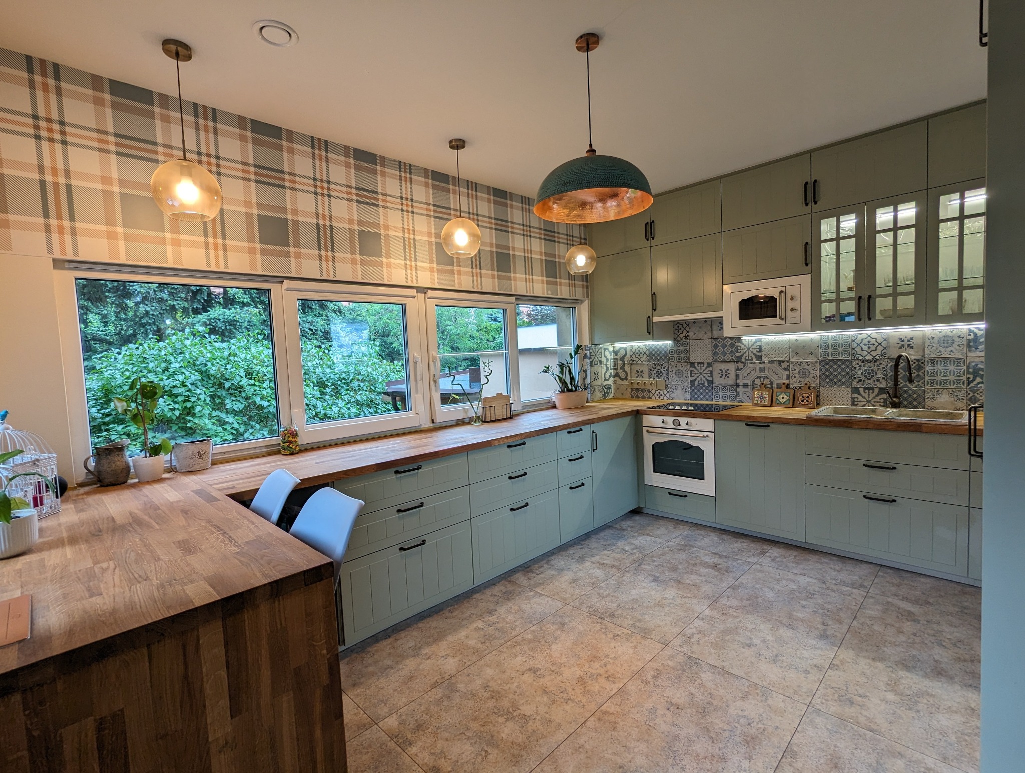 Jasna kuchnia z zielonymi szafkami, drewnianym blatem i wzorzystą tapetą w kratę; okna z widokiem na zieleń, wiszące lampy z bursztynowymi i miedzianymi kloszami.