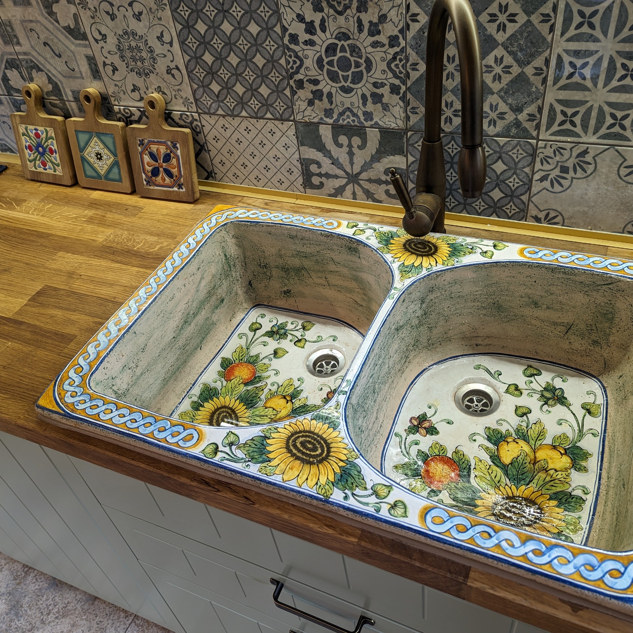 Dwukomorowy zlewozmywak ceramiczny z motywem słoneczników i owoców, osadzony w drewnianym blacie, z brązową baterią i dekoracyjnymi płytkami w tle.