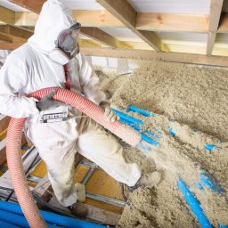 Usługi remontowo budowlane Tomasz Muła - Pracownik w kombinezonie ochronnym aplikuje materiał izolacyjny za pomocą węża do ocieplenia poddasza, widoczne niebieskie rury instalacyjne.