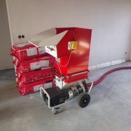Usługi remontowo budowlane Tomasz Muła - Czerwona maszyna do rozdmuchiwania materiału izolacyjnego stoi obok stosu czerwonych paczek wełny mineralnej Rockwool w surowym wnętrzu budynku w trakcie prac ociepleniowych.