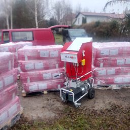 Usługi remontowo budowlane Tomasz Muła - Pakiety wełny mineralnej Rockwool na paletach obok czerwonej maszyny do rozdrabniania i wdmuchiwania izolacji, z czerwonym vanem i domem w tle.