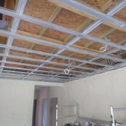 Usługi remontowo budowlane Tomasz Muła - Metalowa konstrukcja sufitu podwieszanego z widocznymi przewodami elektrycznymi, zamontowana pod drewnianym stropem, ściany w trakcie wykańczania, drabina aluminiowa w pomieszczeniu.