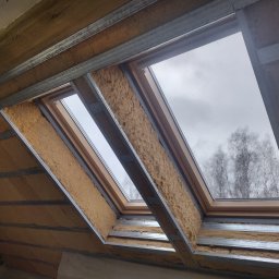 Usługi remontowo budowlane Tomasz Muła - Montaż wełny mineralnej między krokwiami poddasza, widoczne metalowe profile i okna dachowe z widokiem na pochmurne niebo i drzewa.