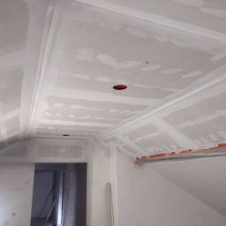 Usługi remontowo budowlane Tomasz Muła - Poddasze w trakcie prac wykończeniowych: widoczne płyty gipsowo-kartonowe na skosach i suficie, częściowo pokryte szpachlą, z wyciętymi otworami na oświetlenie i zabezpieczonymi elementami przed...