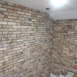 Usługi remontowo budowlane Tomasz Muła - Wykończona ściana wewnętrzna z dekoracyjnej cegły w odcieniach beżu i brązu, widoczna lampa sufitowa, jasne fugi.
