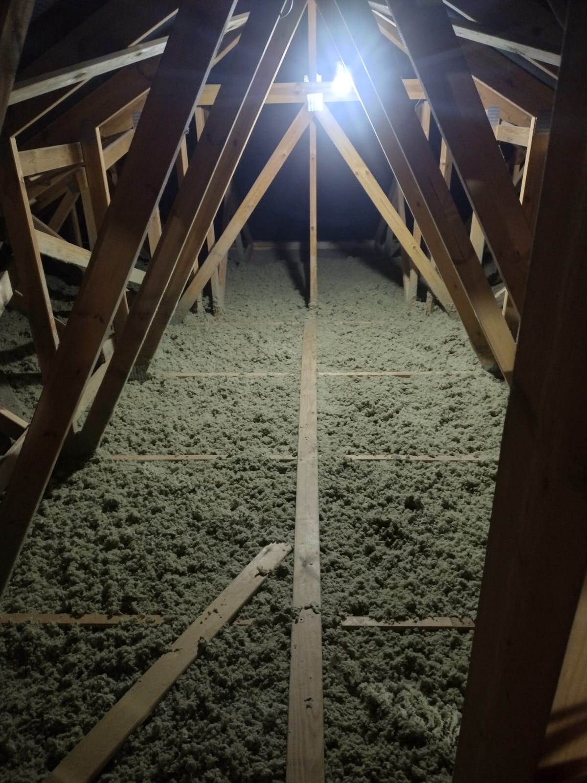 Poddasze z drewnianą konstrukcją wypełnione szarozieloną izolacją, widok z dołu na oświetlenie sufitowe.