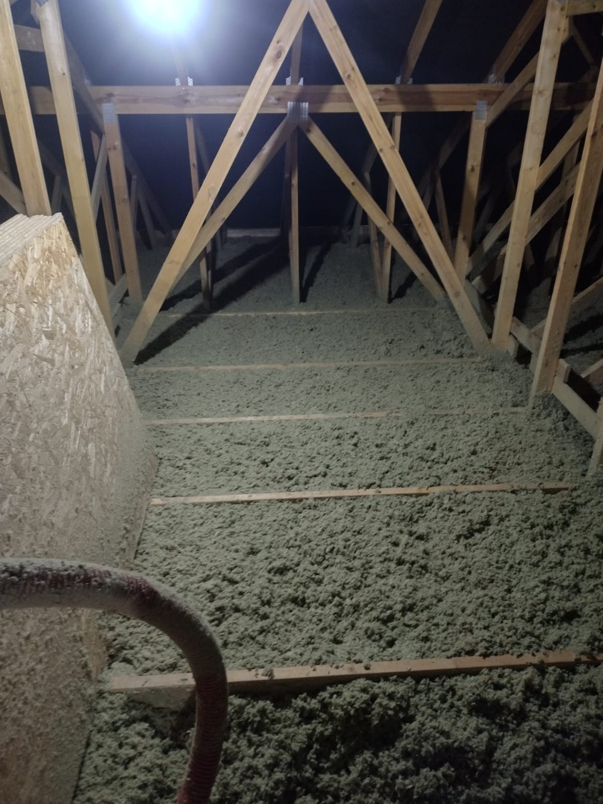 Poddasze z drewnianą konstrukcją pokryte warstwą jasnej izolacji, widoczny fragment węża do wdmuchiwania materiału izolacyjnego i płyta OSB.