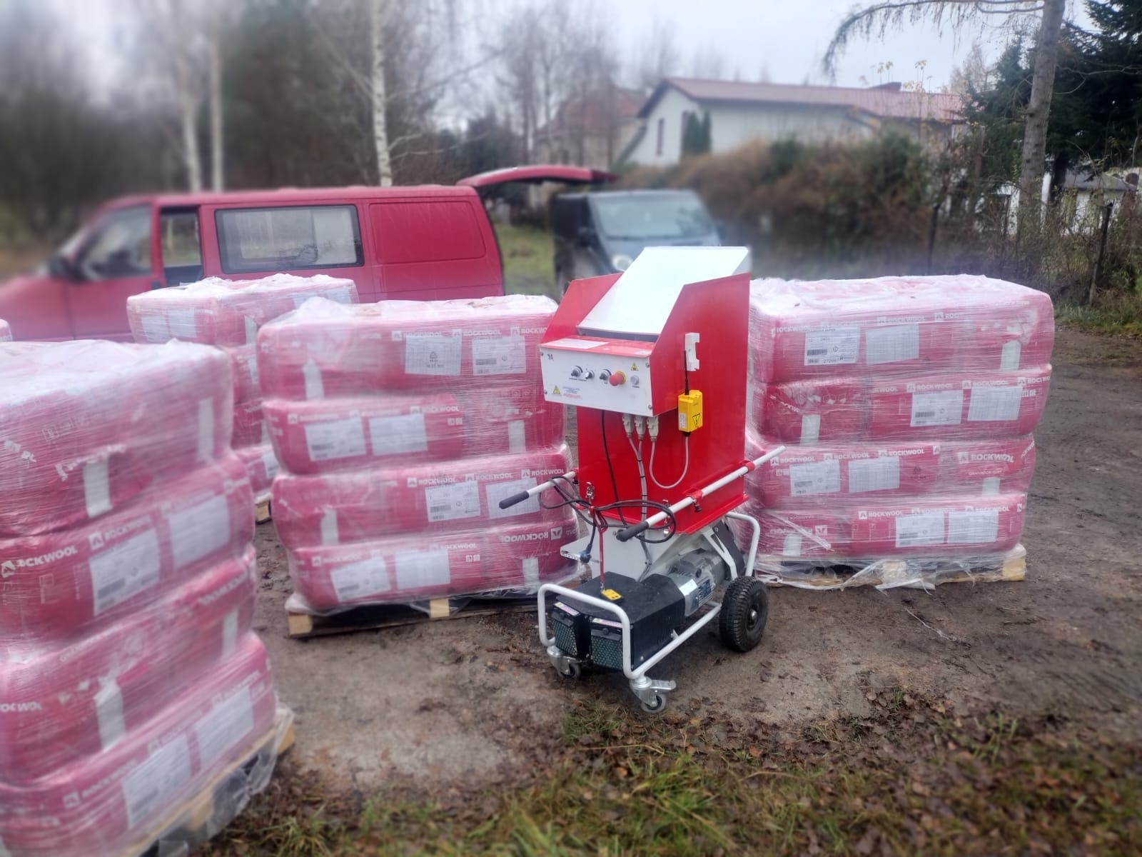 Pakiety wełny mineralnej Rockwool na paletach obok czerwonej maszyny do rozdrabniania i wdmuchiwania izolacji, z czerwonym vanem i domem w tle.