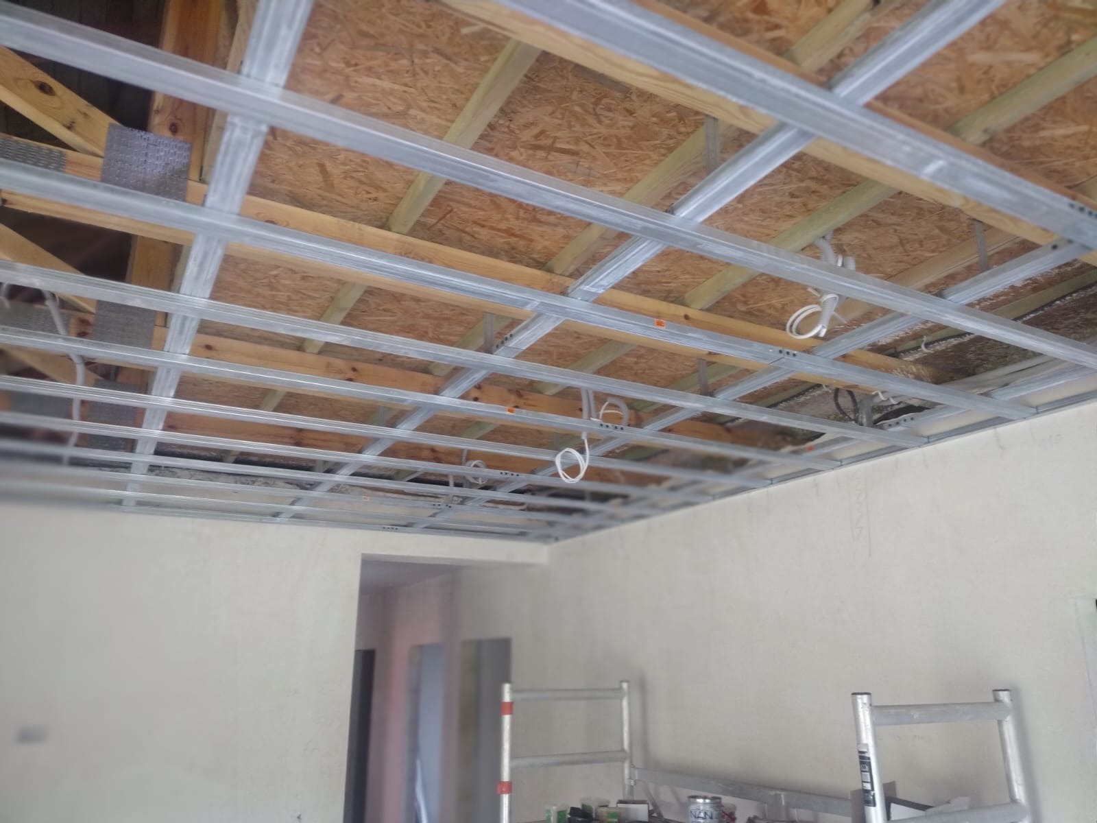 Metalowa konstrukcja sufitu podwieszanego z widocznymi przewodami elektrycznymi, zamontowana pod drewnianym stropem, ściany w trakcie wykańczania, drabina aluminiowa w pomieszczeniu.