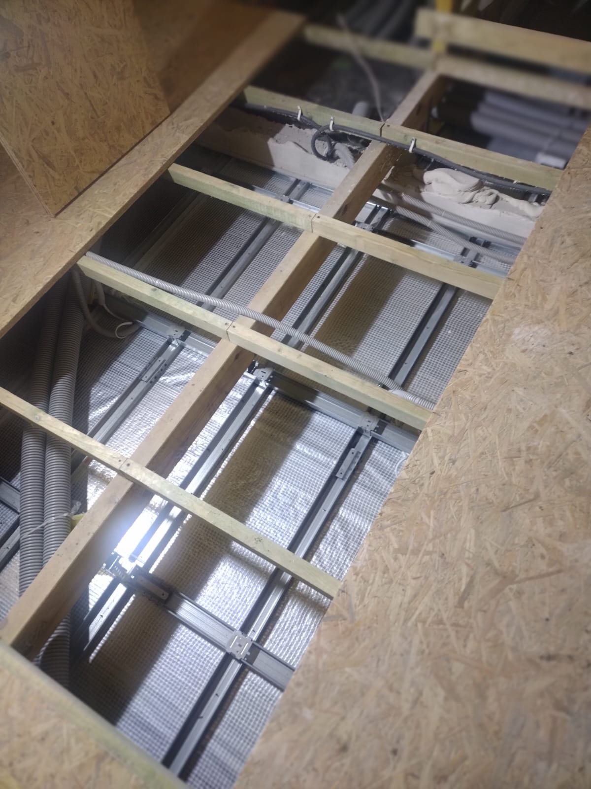 Poddasze w trakcie ocieplania: widoczne drewniane belki, metalowe profile, folia paroizolacyjna i rury wentylacyjne.