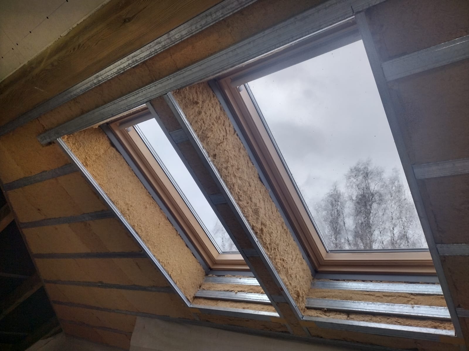 Montaż wełny mineralnej między krokwiami poddasza, widoczne metalowe profile i okna dachowe z widokiem na pochmurne niebo i drzewa.