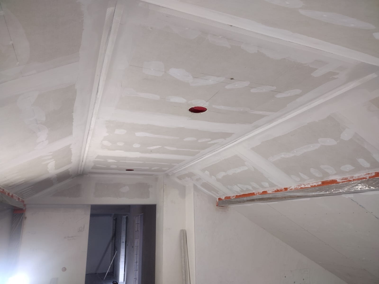 Poddasze w trakcie prac wykończeniowych: widoczne płyty gipsowo-kartonowe na skosach i suficie, częściowo pokryte szpachlą, z wyciętymi otworami na oświetlenie i zabezpieczonymi elementami przed...