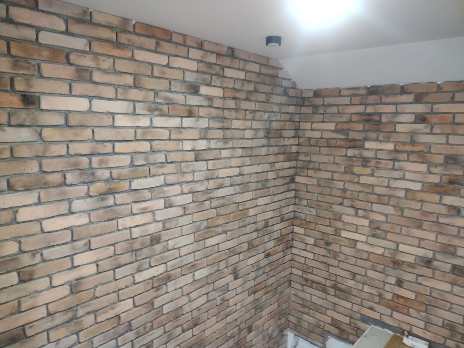 Wykończona ściana wewnętrzna z dekoracyjnej cegły w odcieniach beżu i brązu, widoczna lampa sufitowa, jasne fugi.