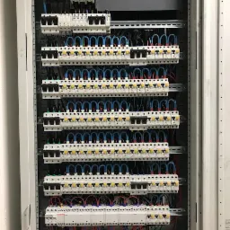 Rozdzielnia elektryczna z licznymi zabezpieczeniami, ułożone rzędami, z niebieskimi i czerwonymi przewodami, widok z przodu, oświetlenie górne.