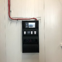 ZPIM.M - Czarna centrala alarmowa z ekranem dotykowym, zamontowana na białej panelowej ścianie, z widocznymi czerwonymi przewodami w korytku kablowym.