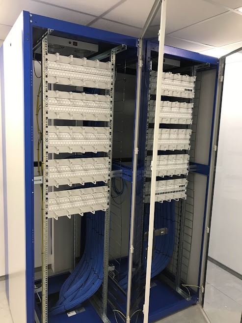 Otwarta szafa serwerowa w kolorze niebiesko-białym, zawierająca panele krosownicze z dużą ilością niebieskich kabli sieciowych uporządkowanych w wiązki.