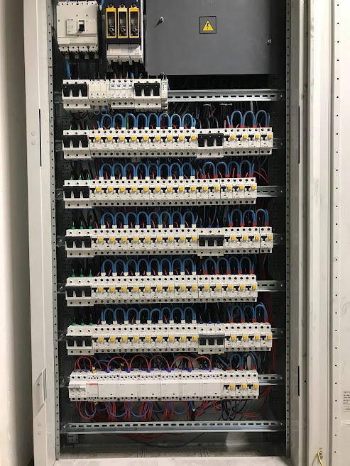 Rozdzielnia elektryczna z licznymi zabezpieczeniami, ułożone rzędami, z niebieskimi i czerwonymi przewodami, widok z przodu, oświetlenie górne.