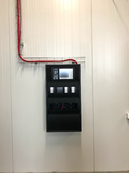 Czarna centrala alarmowa z ekranem dotykowym, zamontowana na białej panelowej ścianie, z widocznymi czerwonymi przewodami w korytku kablowym.