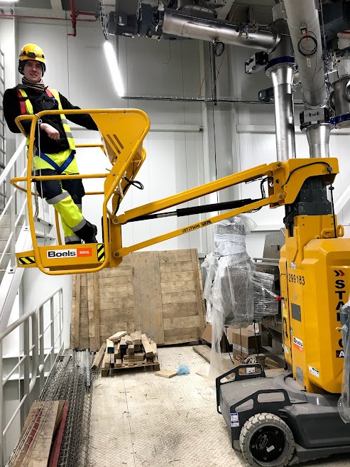 Pracownik w żółtym kasku na podnośniku koszowym Boels w hali w Warszawie, prawdopodobnie podczas montażu instalacji przemysłowej, widoczne rury i skrzynie.