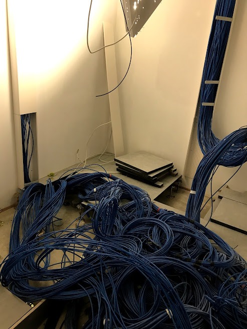 Stos niebieskich kabli ethernetowych na podłodze w trakcie instalacji, z widocznymi otworami w ścianie i korytkami kablowymi.