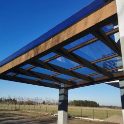 Aleksander Halczuk - Drewniana pergola z przeszklonym dachem, widok z dołu na błękitne niebo. Konstrukcja wsparta na szarych słupach, w tle zielone pole i drzewa.