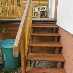 Schody z drewna prowadzące na taras z ażurową ścianą. Widoczny zielony pojemnik i krzesło na tarasie. Ciepłe oświetlenie podkreśla strukturę drewna.