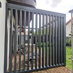 Aleksander Halczuk - Nowoczesna, szara pergola z pionowych desek na tarasie z kostki. Widok przez szczeble na meble ogrodowe i zieleń. Minimalistyczny design.