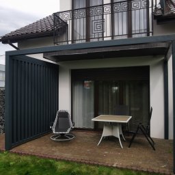 Aleksander Halczuk - Nowoczesna pergola z metalową konstrukcją, częściowo zabudowana, z meblami ogrodowymi na ceglanym tarasie. Widok na dom z balkonem z ozdobną balustradą.