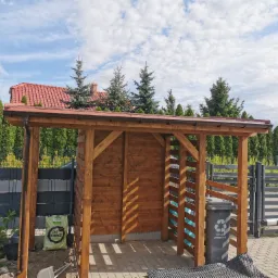 Drewniana konstrukcja z zadaszeniem, prawdopodobnie wiata śmietnikowa, z brązowym dachem i ścianami, na tle błękitnego nieba i zielonych drzew.