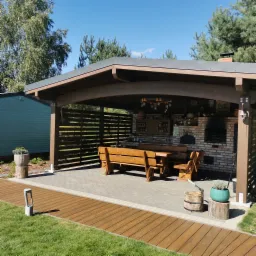 Zadaszona altana z ceglanym grillem i drewnianym stołem. Elementy drewniane w ciepłym brązie, na zewnątrz trawnik i błękitne niebo. Widoczny fragment drewnianego chodnika.