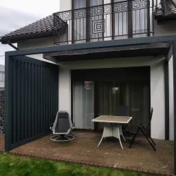 Nowoczesna, grafitowa pergola z metalu przy białym domu. Na tarasie stół i krzesła. Balustrada balkonu z geometrycznym wzorem.