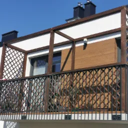 Nowoczesny balkon z metalową balustradą i drewnianą konstrukcją kratową, uzupełniony roślinami w donicach, na tle błękitnego nieba.