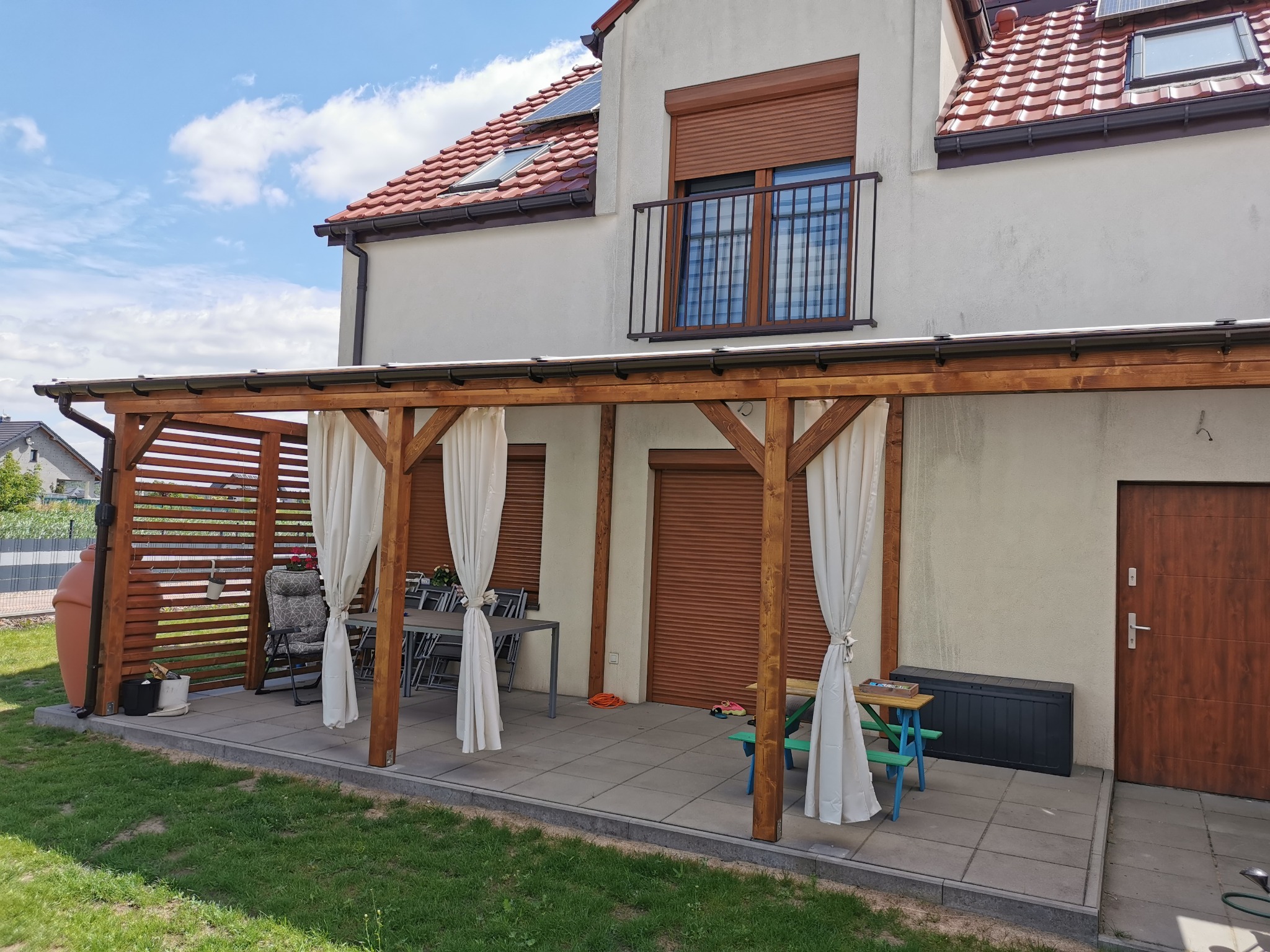 Drewniana pergola z białymi zasłonami na tarasie w Brzegu Dolnym, przylegająca do domu z balkonem i roletami, tworząca zacienioną przestrzeń wypoczynkową.