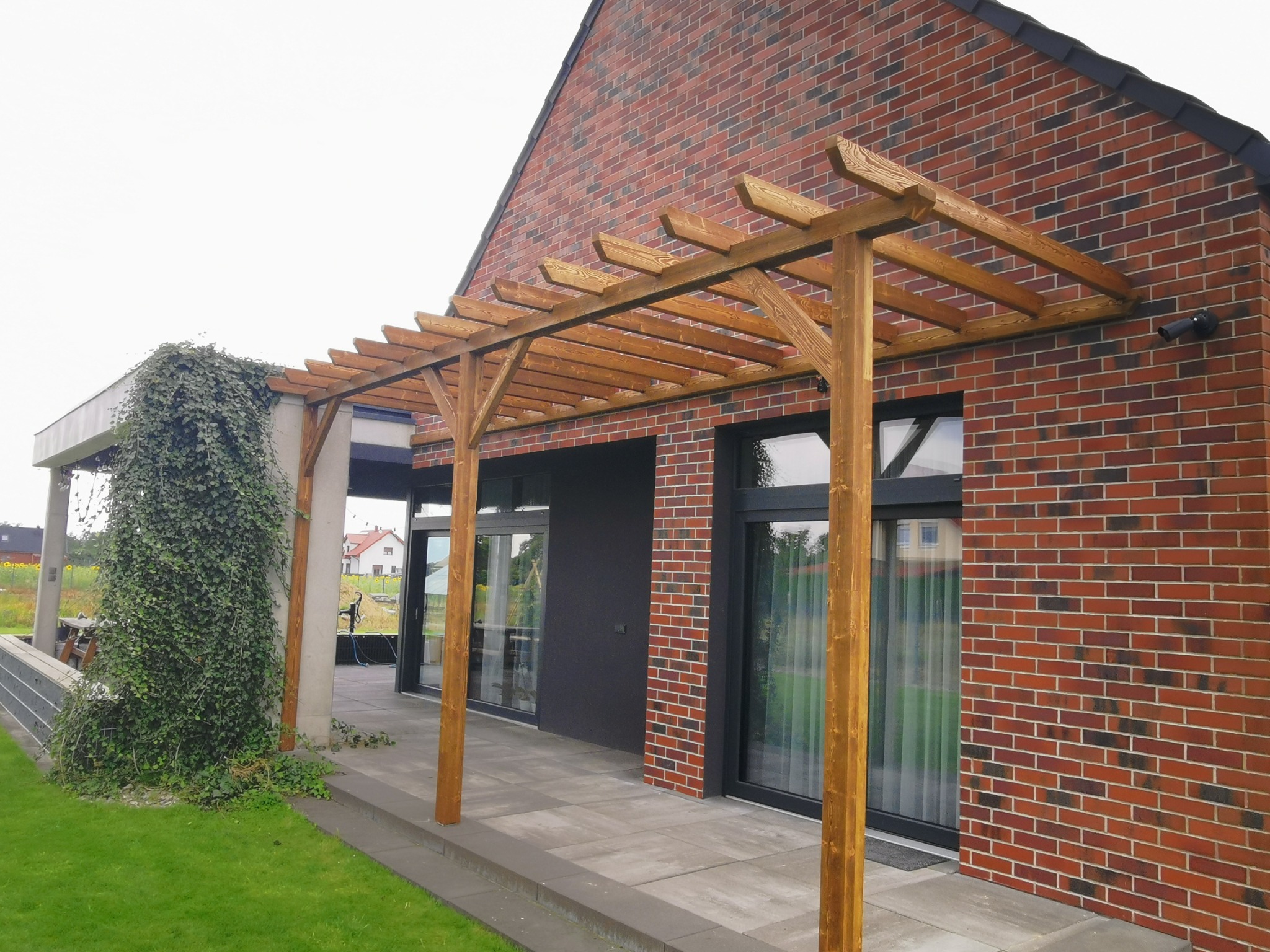 Drewniana pergola z belek nad tarasem z szarych płyt, przylegająca do ceglanego domu z dużymi oknami, zarośnięta pnączem po lewej stronie.