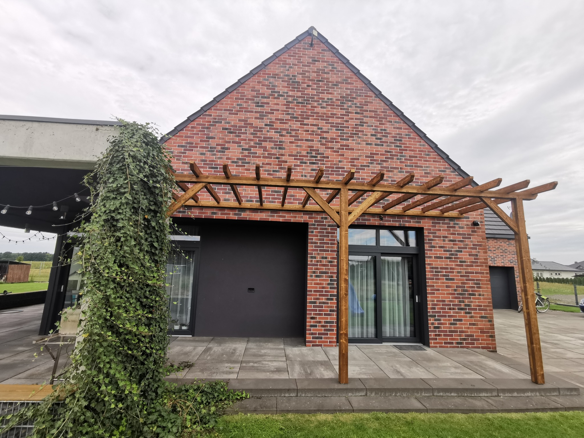 Nowoczesna pergola drewniana przy ceglanym domu z szarym tarasem, obrośnięta bluszczem. Minimalistyczny design, naturalne materiały, pochmurne niebo w tle.