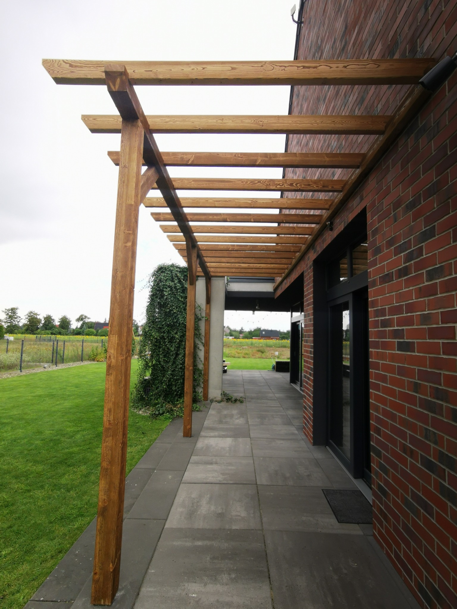 Drewniana pergola przy ceglanym budynku, z widokiem na trawnik. Konstrukcja z belek, szara kostka brukowa na podłożu, zieleń pnąca się po filarze.