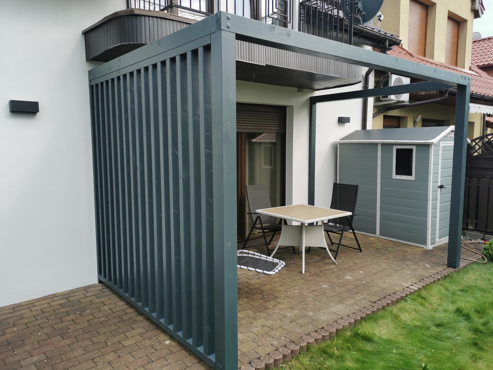 Nowoczesna, szara pergola z pionowymi listwami, zadaszona częściowo, z meblami ogrodowymi i domkiem narzędziowym na brukowanym tarasie.