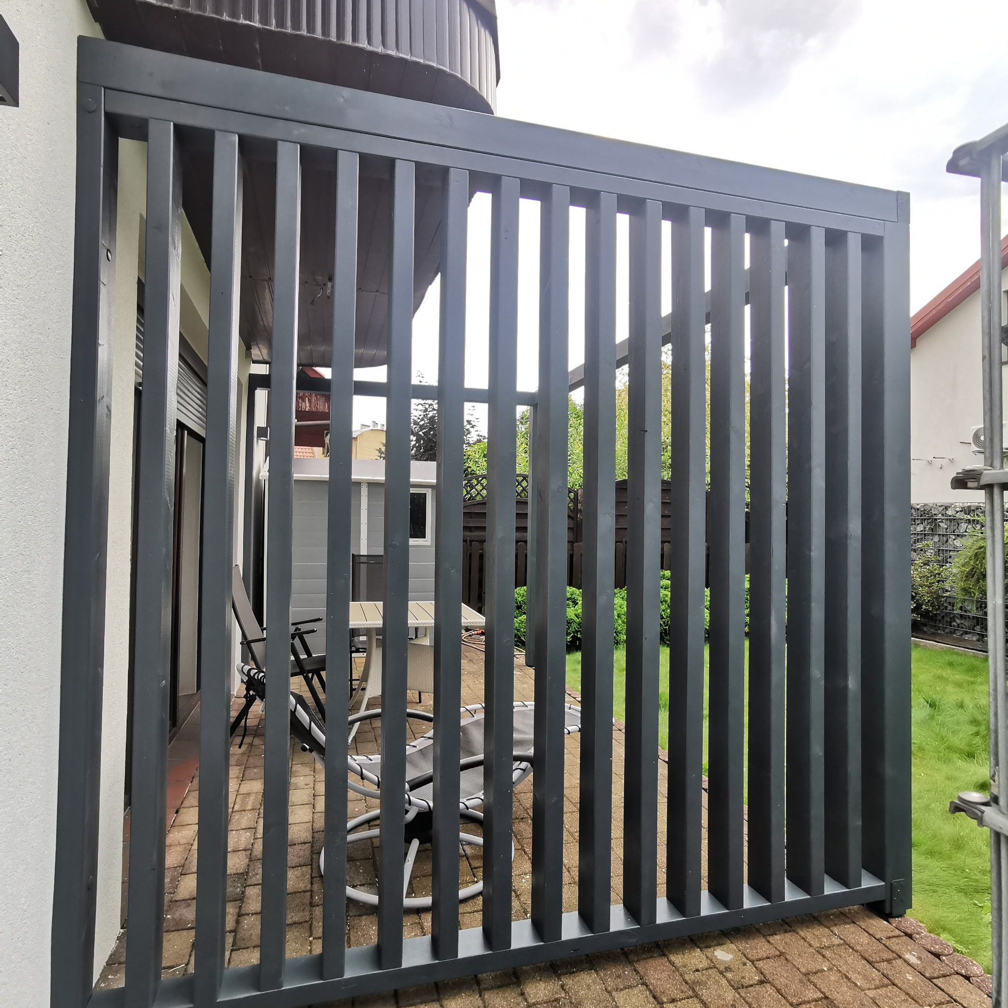 Nowoczesna, szara pergola z pionowych desek na tarasie z kostki. Widok przez szczeble na meble ogrodowe i zieleń. Minimalistyczny design.