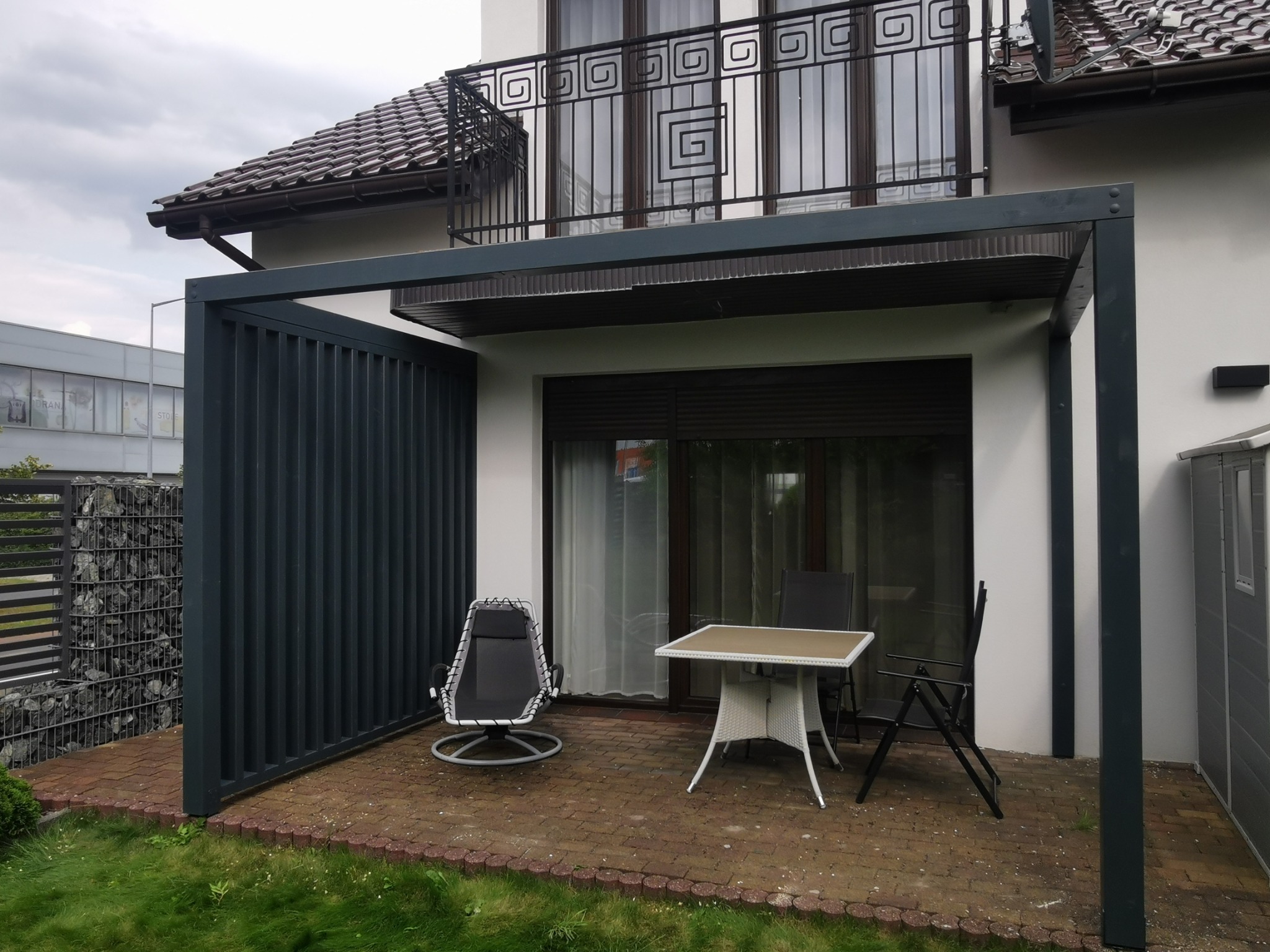 Nowoczesna pergola z metalową konstrukcją, częściowo zabudowana, z meblami ogrodowymi na ceglanym tarasie. Widok na dom z balkonem z ozdobną balustradą.