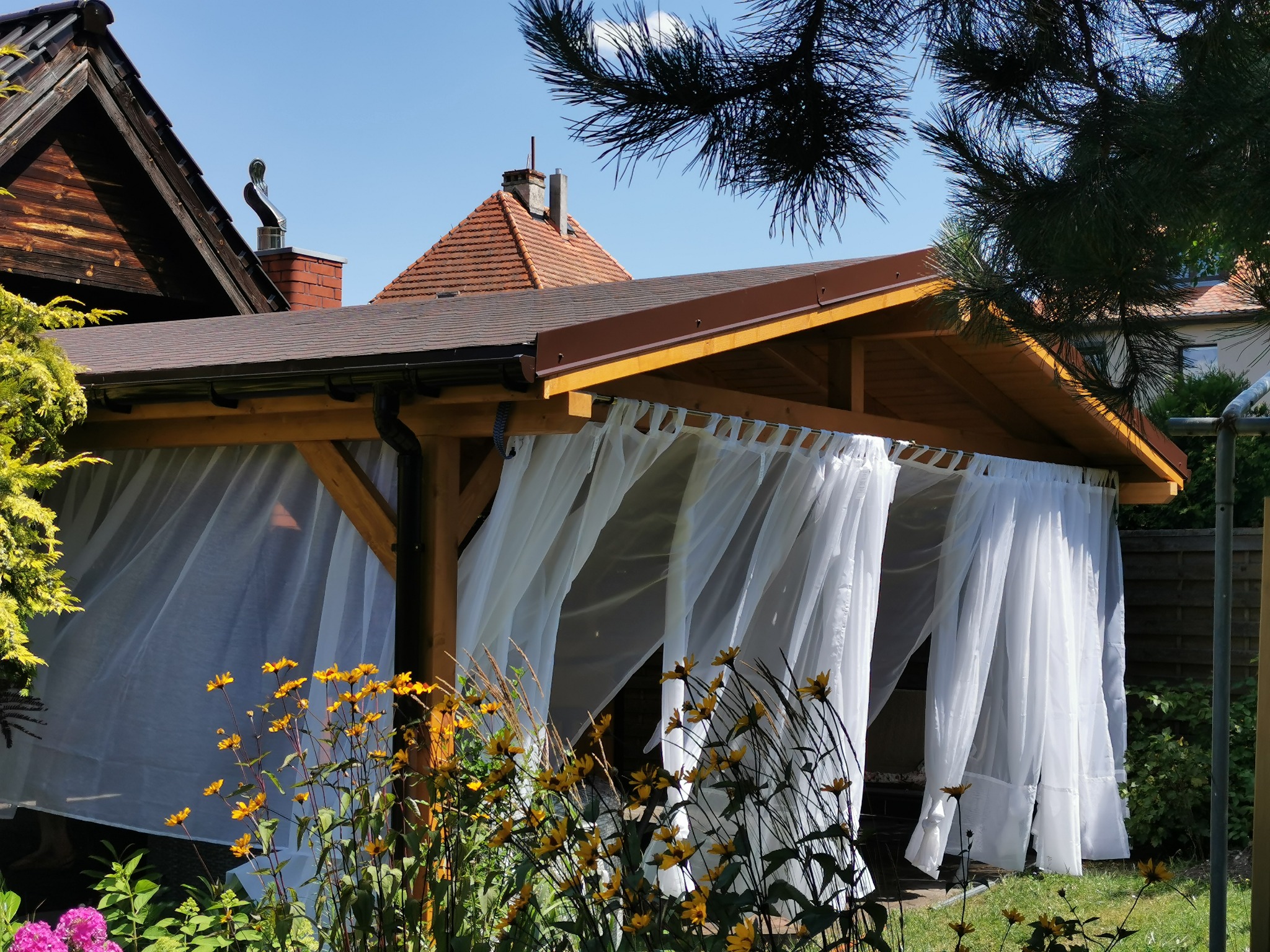 Drewniana altana z białymi zasłonami, otoczona kwitnącymi kwiatami w ogrodzie, w tle domy z dachówką, słoneczny dzień.