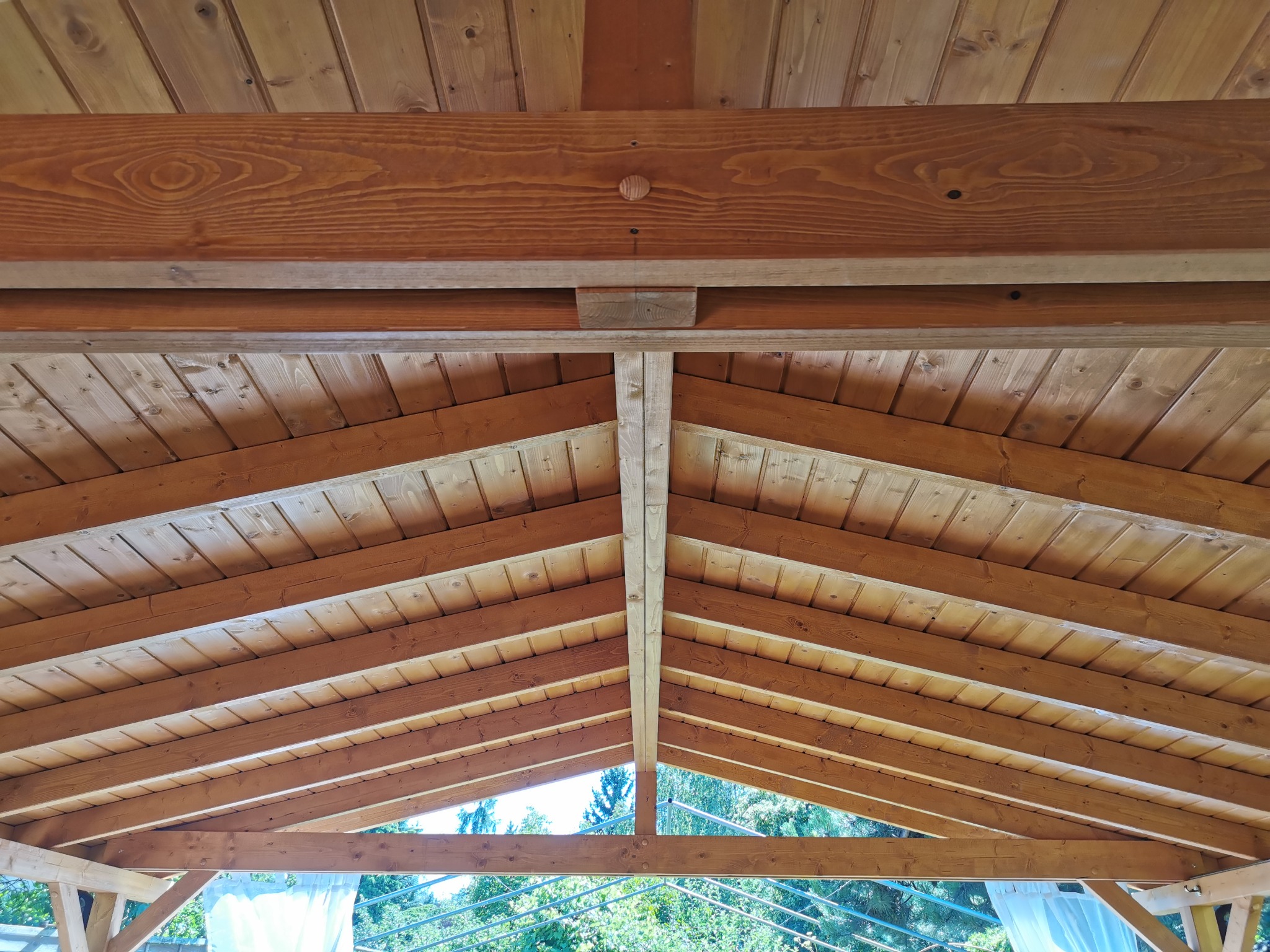 Drewniany szkielet dachu altany z widoczną konstrukcją krokwi, belek stropowych i deskowaniem. Widok z dołu na zewnątrz, zieleń w tle.