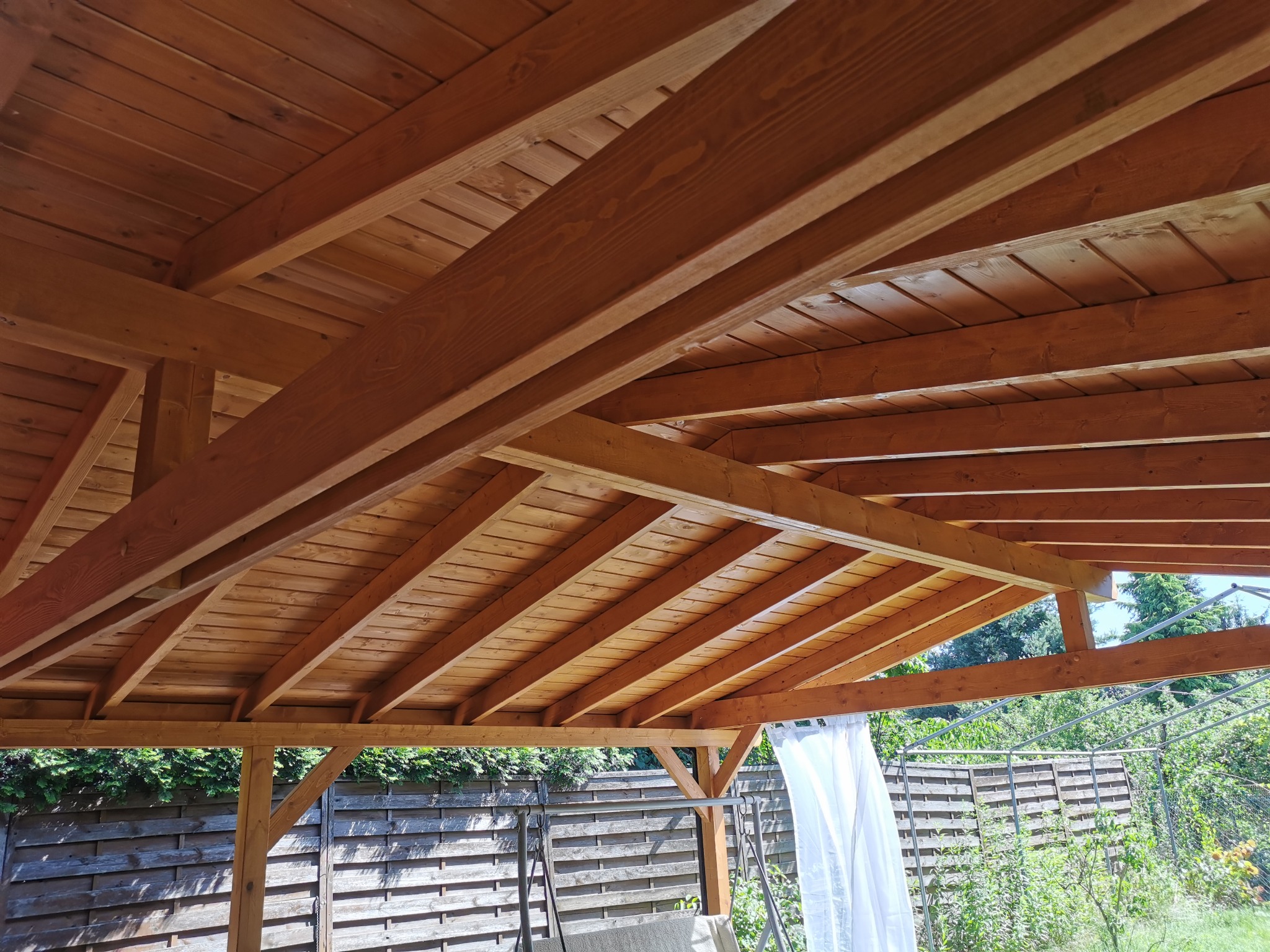 Drewniana konstrukcja z belek i desek, prawdopodobnie altana lub pergola, widziana od dołu. Widoczne elementy ogrodu w tle.