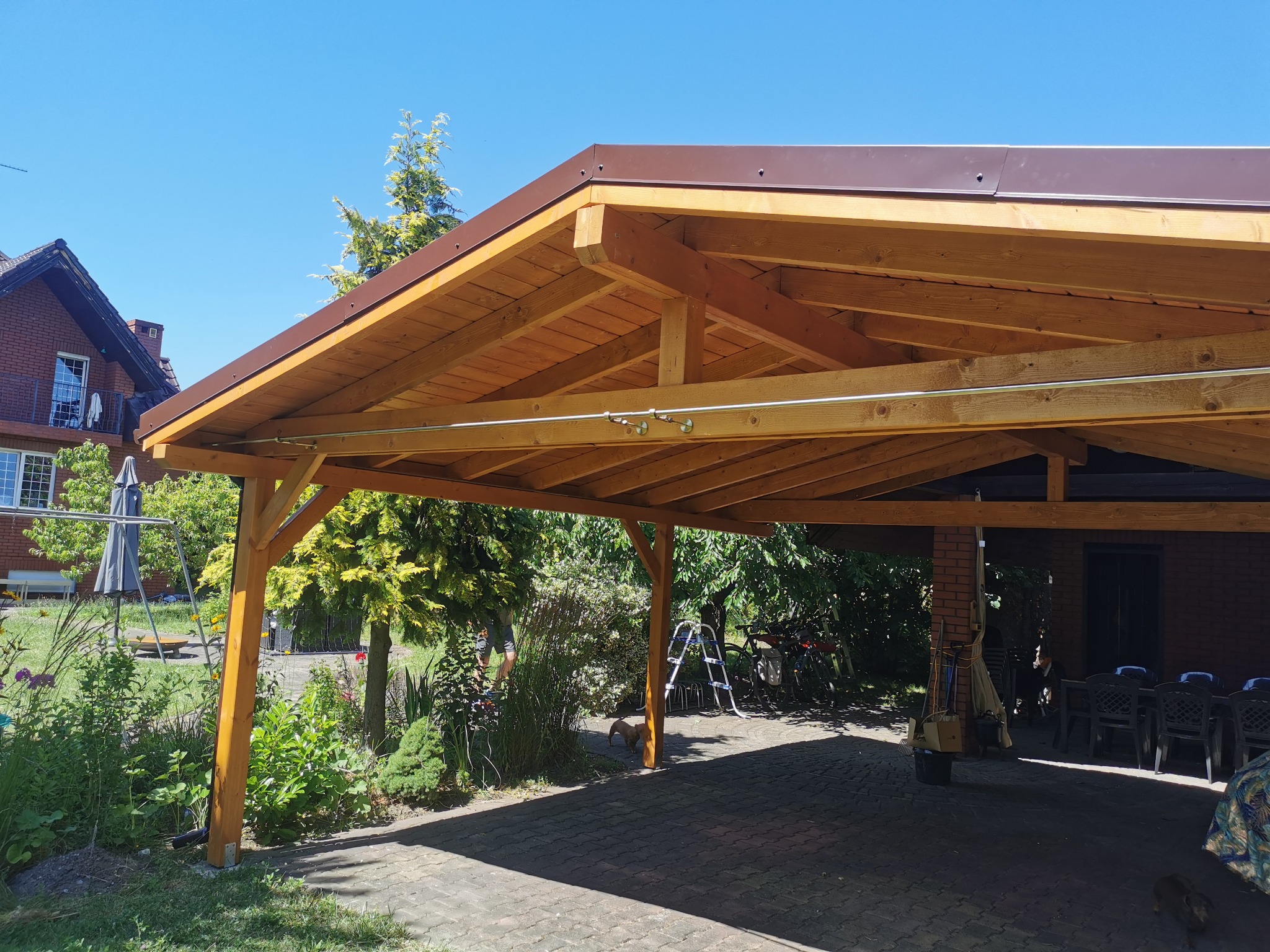 Drewniana wiata z brązowym dachem, wsparta na solidnych słupach, osłaniająca podjazd z kostki. W tle zieleń ogrodu i dom z czerwonej cegły.