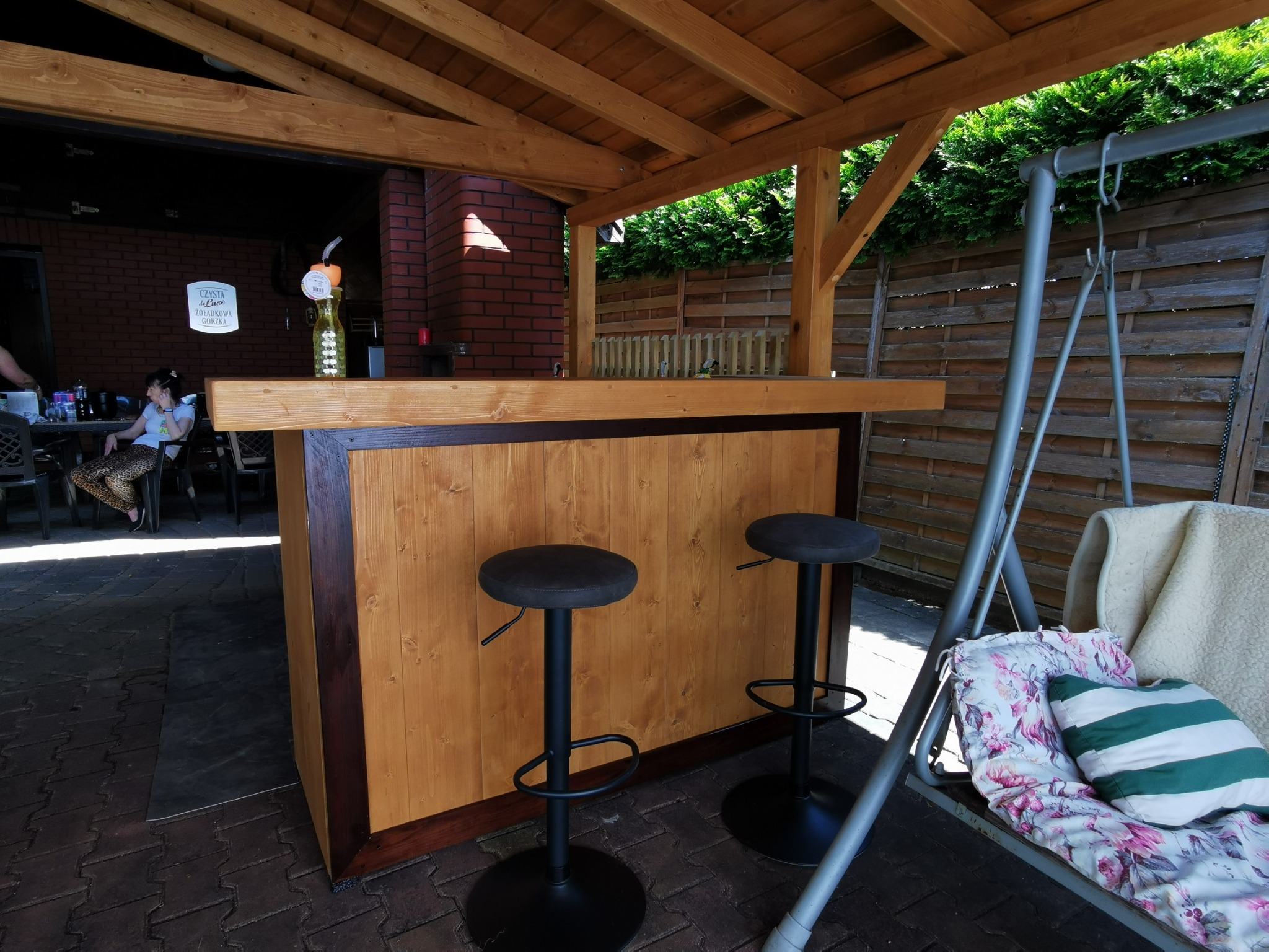 Drewniany bar z dwoma stołkami pod zadaszeniem, obok huśtawka. W tle ceglany budynek i drewniany płot. Oświetlenie naturalne, słoneczny dzień.