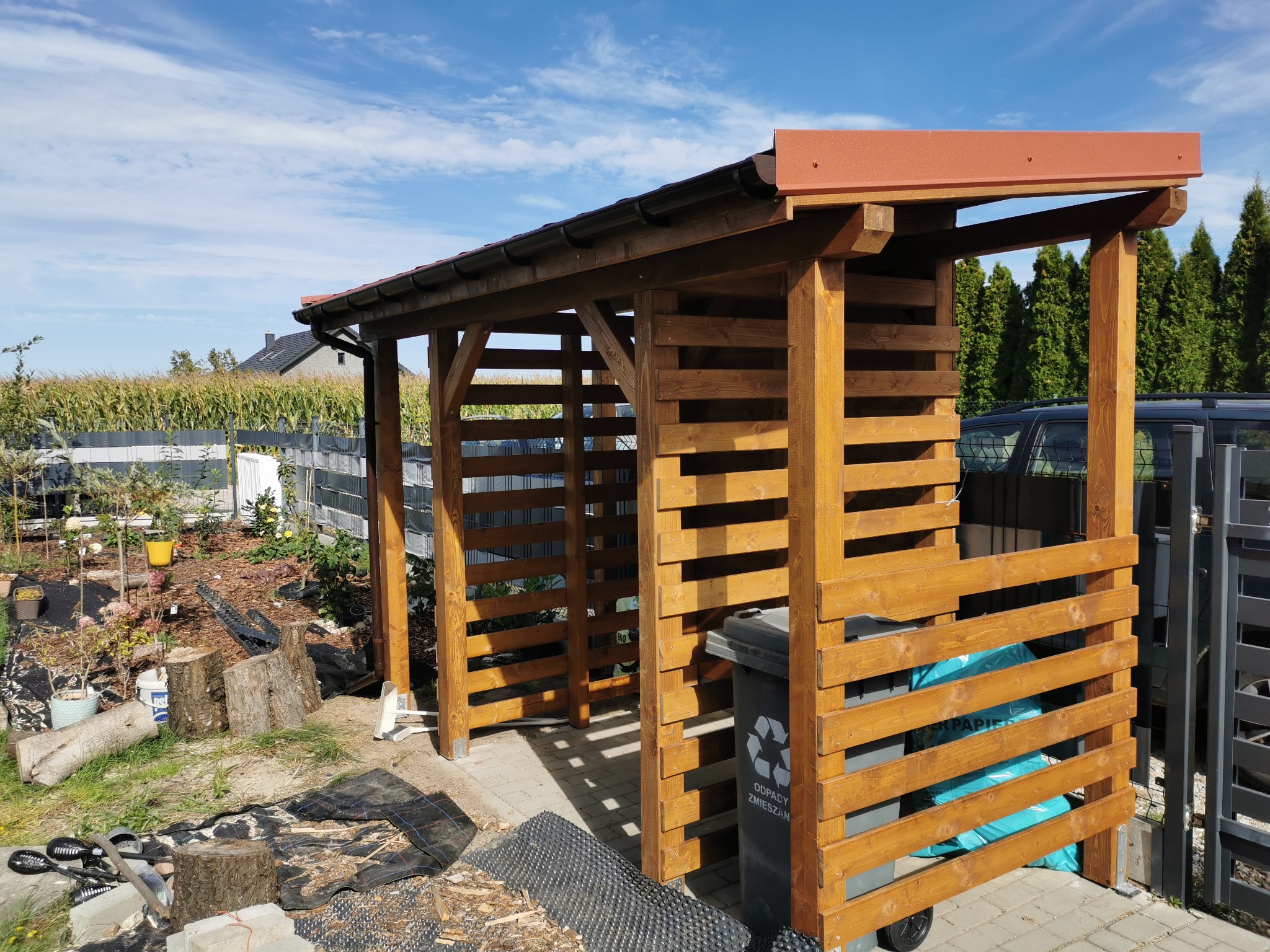 Drewniana wiata na śmietniki z brązowym dachem, z widocznymi pojemnikami na odpady, na tle ogrodu i błękitnego nieba. Konstrukcja z poziomych desek.