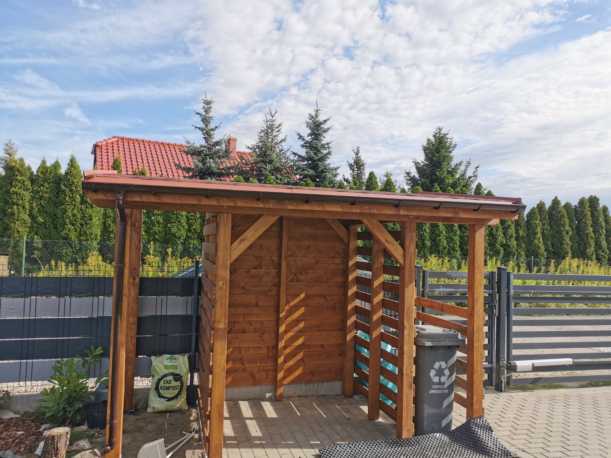 Drewniana konstrukcja z zadaszeniem, prawdopodobnie wiata śmietnikowa, z brązowym dachem i ścianami, na tle błękitnego nieba i zielonych drzew.