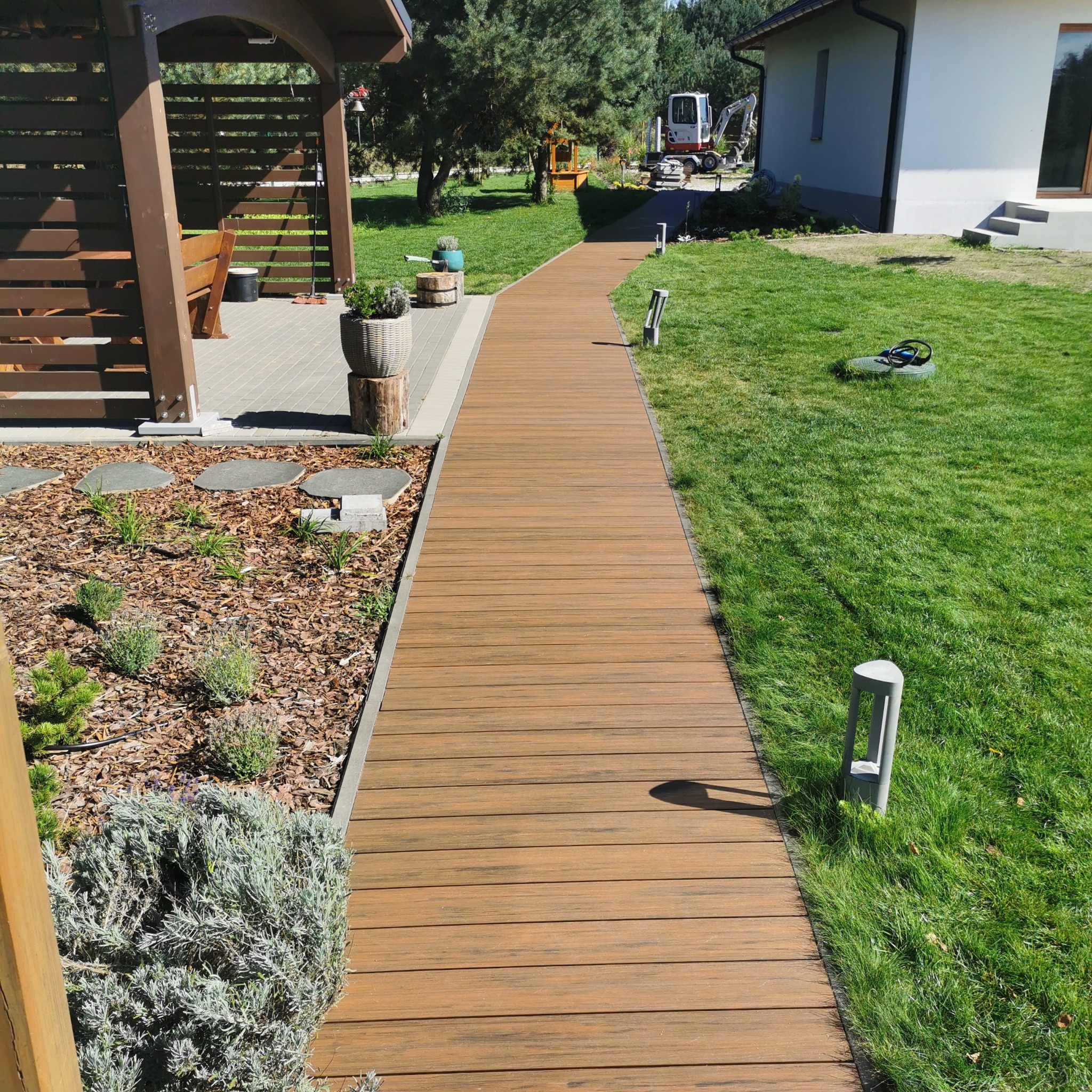 Kompozytowa ścieżka ogrodowa prowadzi od altany do domu, oświetlona lampami. W tle trawnik i maszyna budowlana.