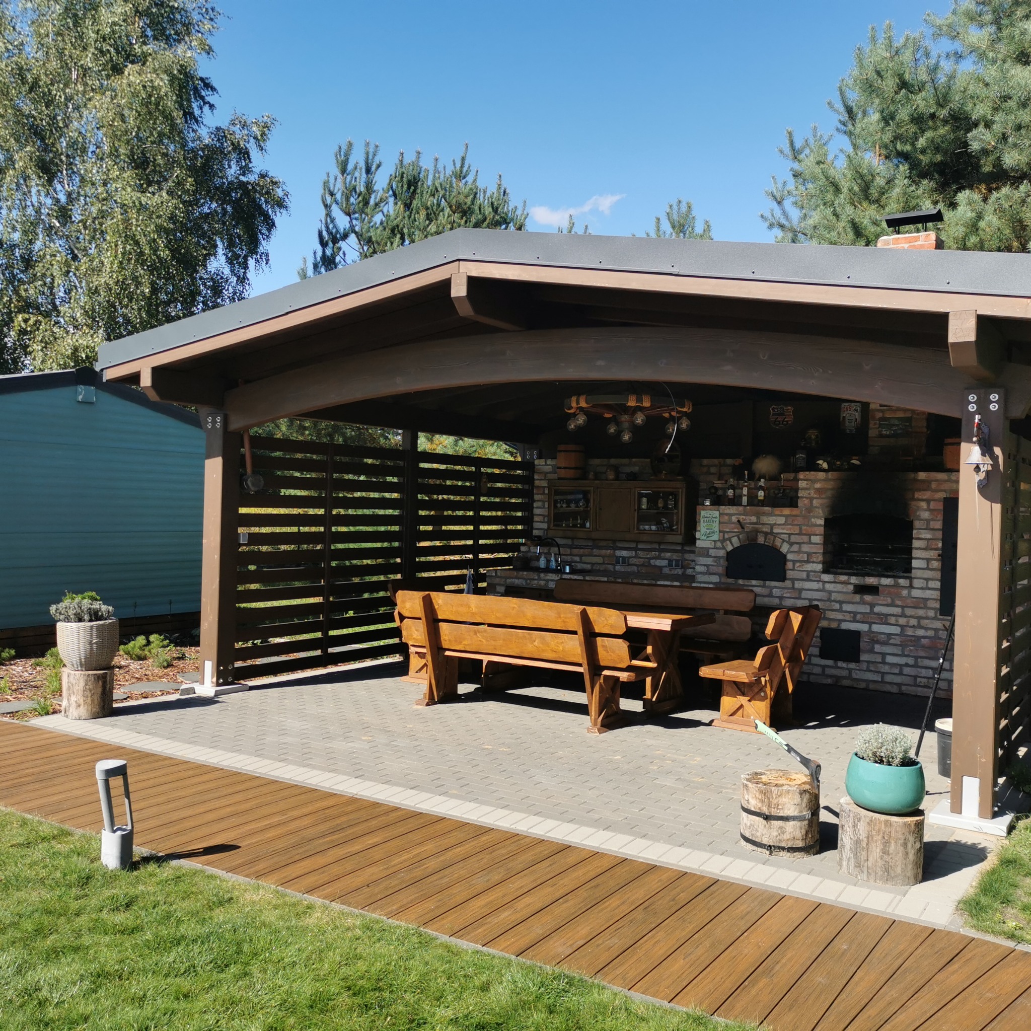 Zadaszona altana z ceglanym grillem i drewnianym stołem. Elementy drewniane w ciepłym brązie, na zewnątrz trawnik i błękitne niebo. Widoczny fragment drewnianego chodnika.