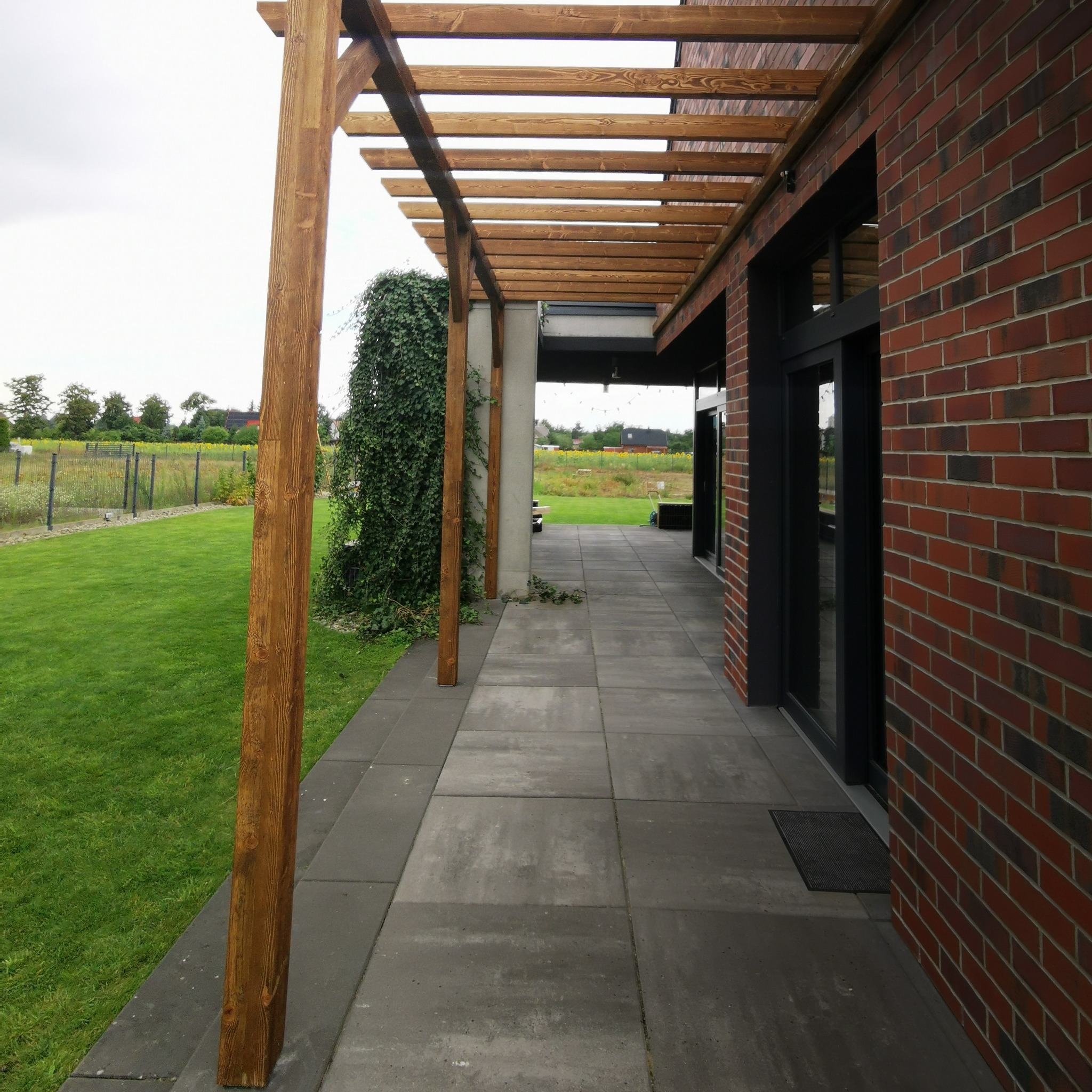 Nowoczesna pergola drewniana z betonową posadzką tarasu, przylegająca do budynku z cegły. Widok na zadbany trawnik i zieleń w tle.